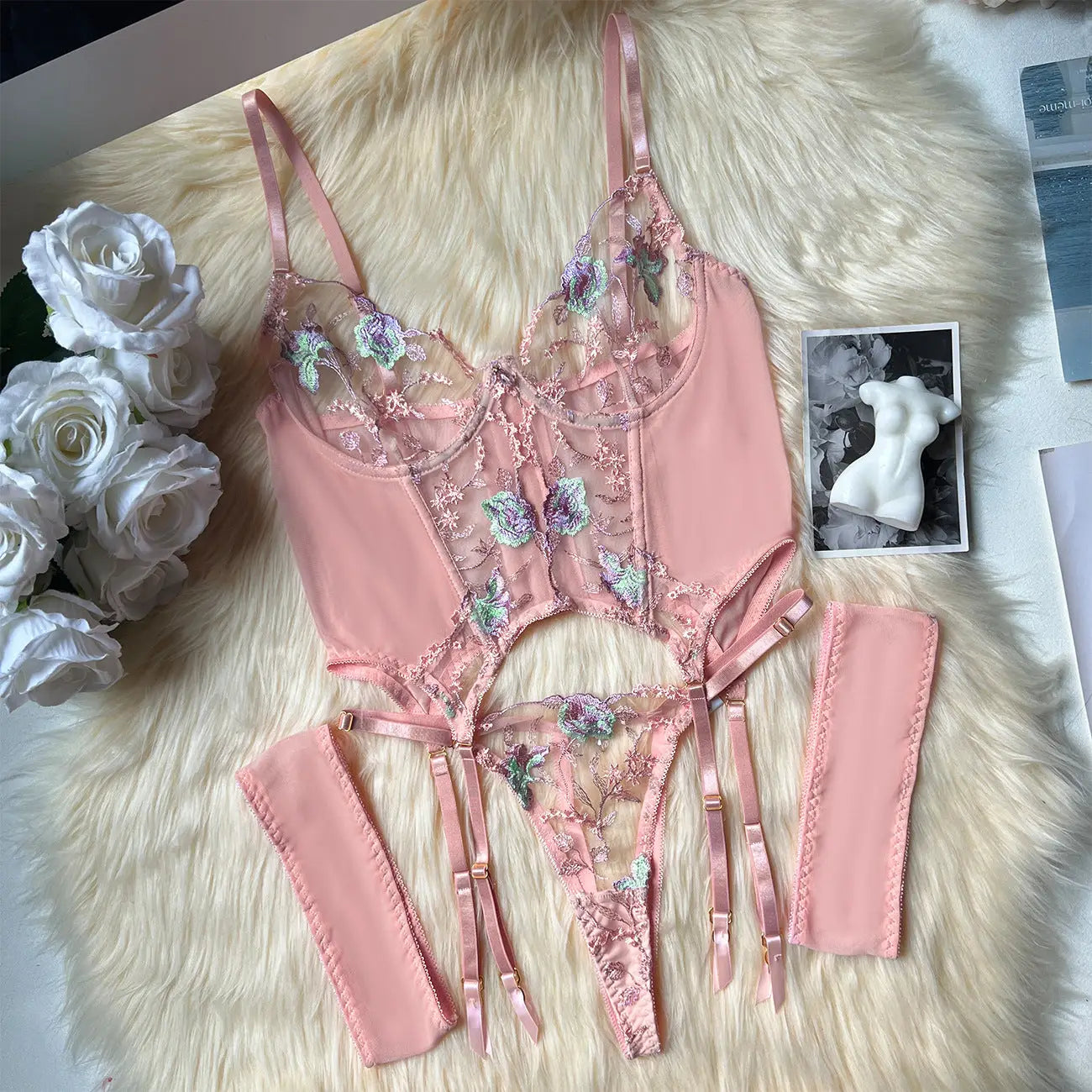 Pink lingerie set with floral embroidery on a fluffy white surface with flowers and a photo frame.