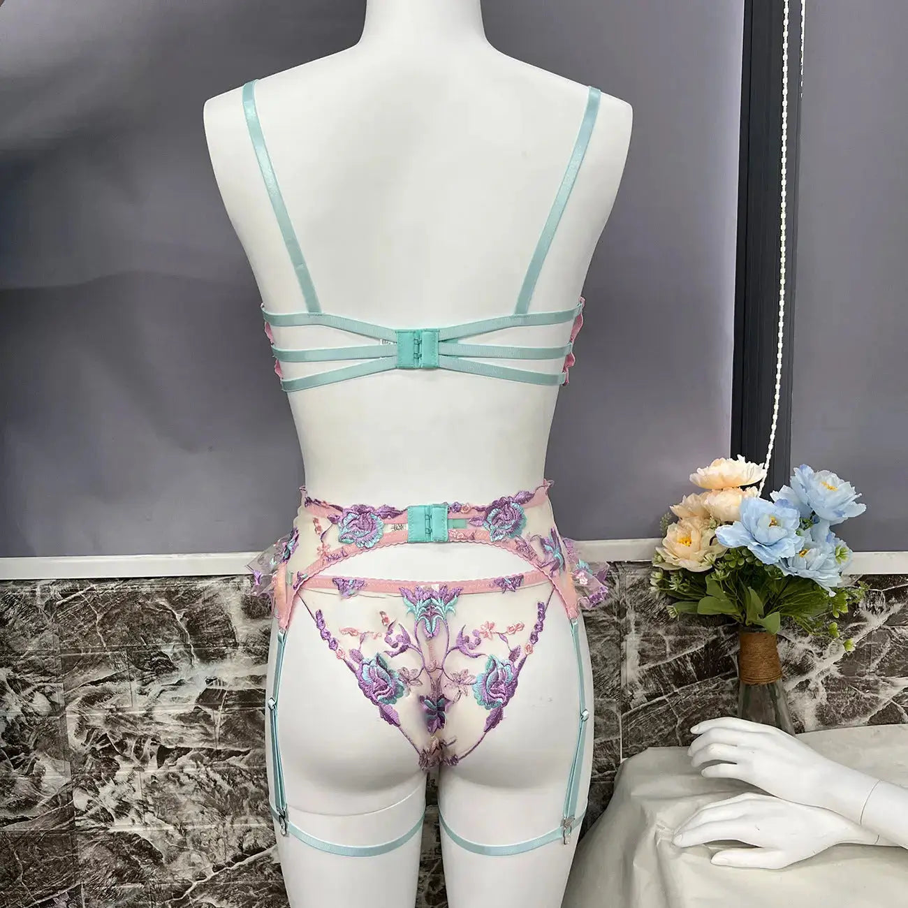 Elegant lingerie set featuring a pastel mint strappy bra, intricately embroidered sheer garter belt, and matching thong with vibrant floral motifs in pink and purple, displayed on a mannequin.