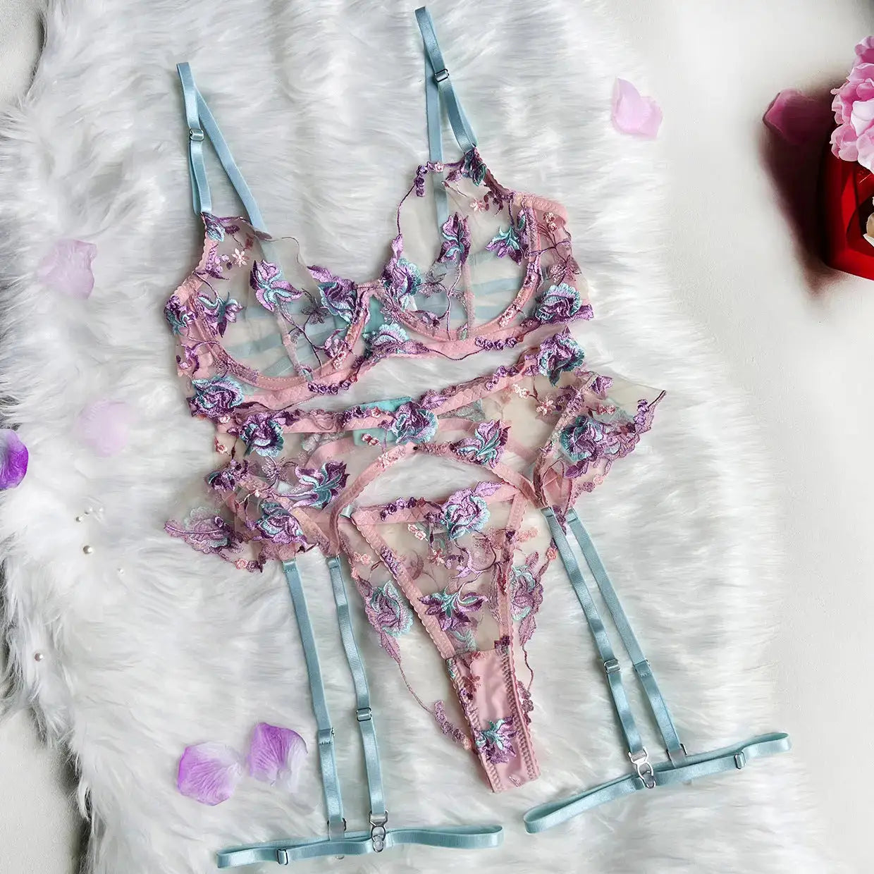 A delicate sheer pink lingerie set, featuring a bra, thong, and garter belt. It's adorned with intricate blue and purple floral embroidery and light blue adjustable straps.