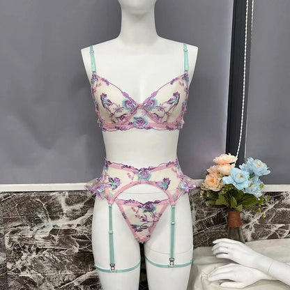 An elegant three-piece lingerie set featuring a bra, garter belt, and thong. It has delicate floral embroidery in purple, teal, and pink on sheer white fabric, accented with mint green straps.